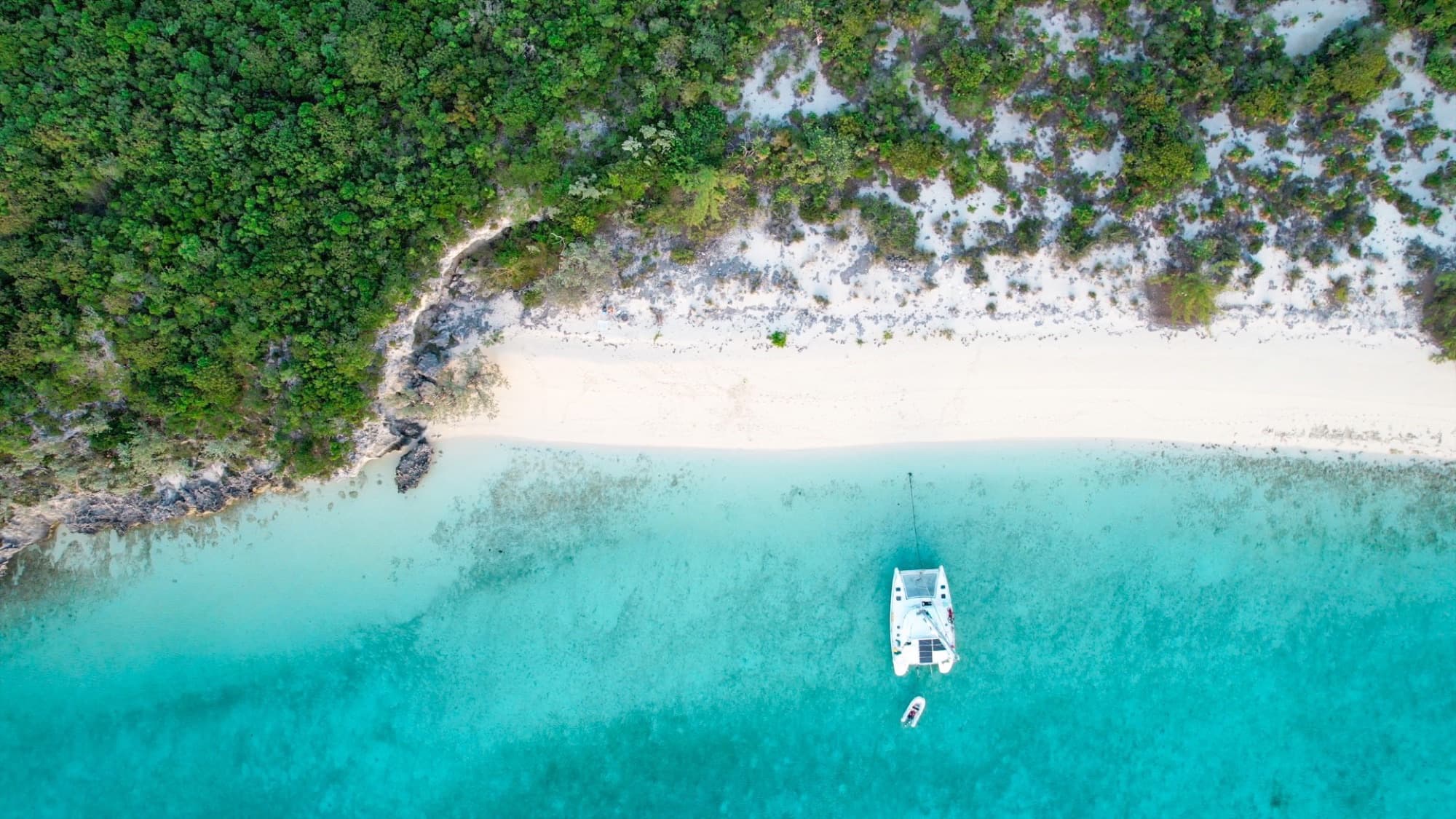 Honu at anchor in turquoise water off a white beach, seen from above